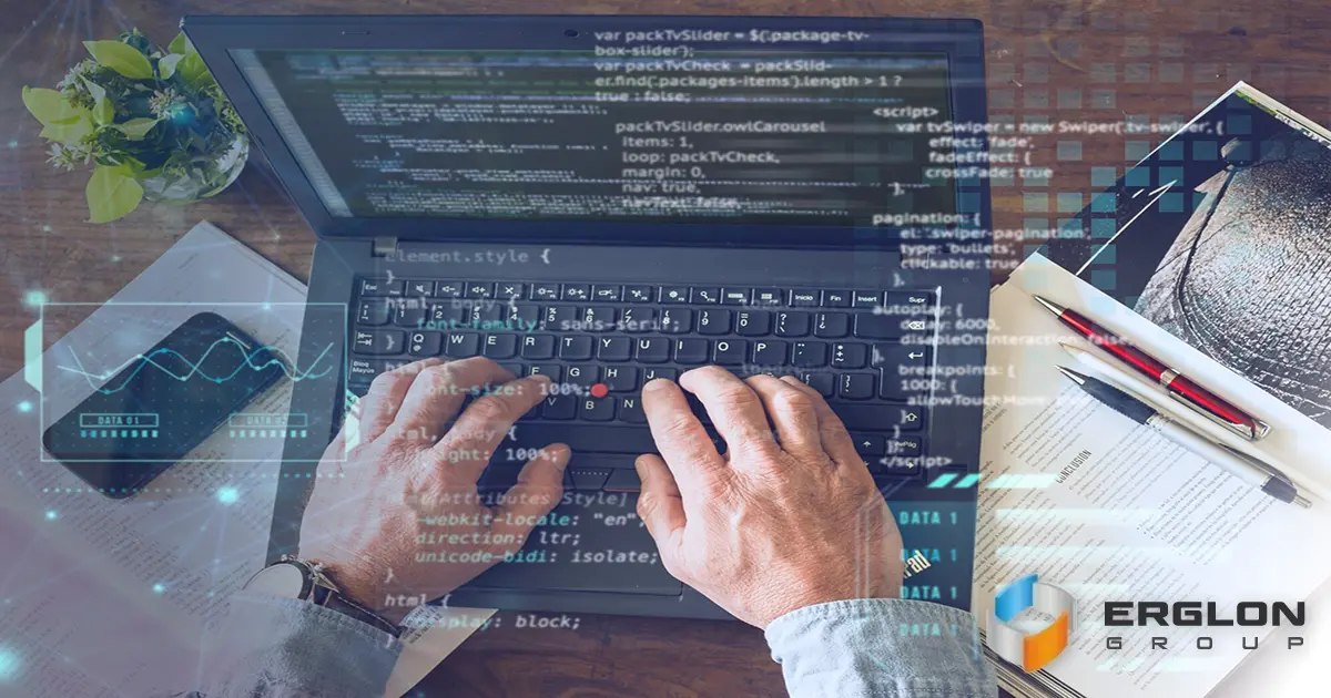 A software developer sitting at a desk typing on a laptop with lines of source code visible on the screen in a modern workspace.